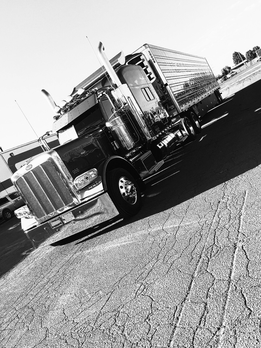 Black-and-white photograph of a semi-truck and trailer on an asphalt lot, shot at a dynamic diagonal angle under bright sunlight